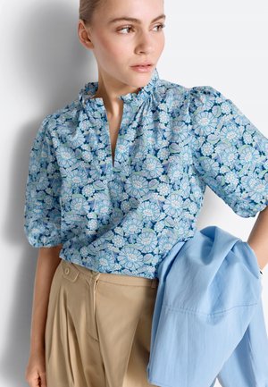 Frau trägt eine blaue Bluse mit Blumenmuster und Puffärmeln sowie beige Hosen, hält eine hellblaue Jacke über ihrem Arm.