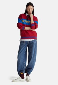 Bluza w czerwono-niebieskie bloki kolorystyczne z logo "BENETTON", zestawiona z luźnymi dżinsami i białymi tenisówkami. Modelka w warstwowym ubraniu.