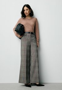 Brauner Pullover, graue karierte weit geschnittene Hose, schwarzer Gürtel und spitze schwarze Schuhe. Hält eine schwarze Tasche. Glatte Texturen und strukturierte Formen.