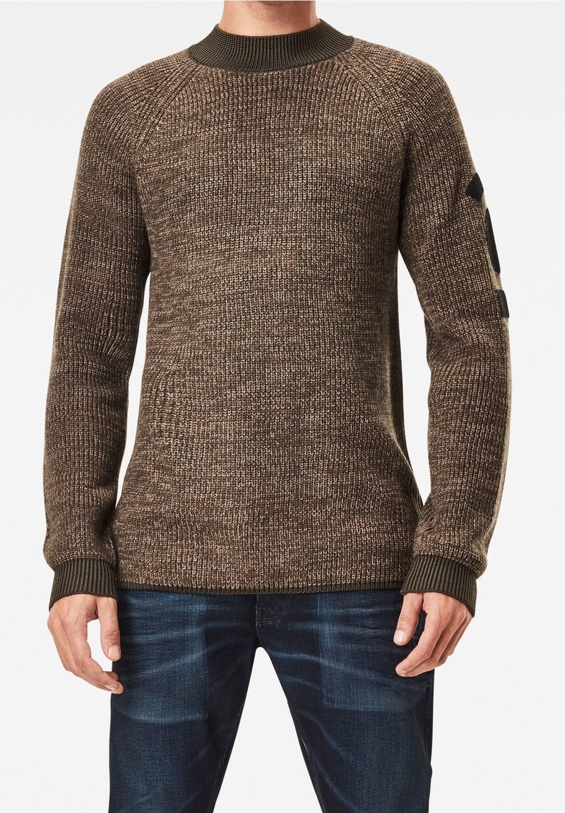 Mann in einem braunen strukturierten Strickpullover mit grünem Rippkragen und -bündchen, kombiniert mit dunkelblauen Jeans.