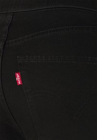 Crne traperice od denima ističu se istaknutom crvenom Levi's oznakom na stražnjem džepu. Tkanina ima glatku teksturu s suptilnim detaljima šivanja.