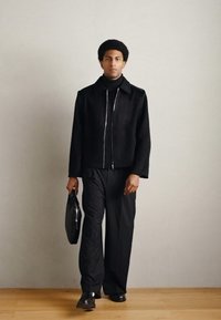 Crna jakna od brušene kože s patentnim zatvaračem, crni rolka džemper, široke crne hlače i sjajne crne cipele. Drži crnu aktovku.