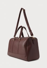 Hnedá kožená taška typu duffel s textúrou kamenistého povrchu, dvojité horné rukoväte a odnímateľný popruh. Zlatej farby kovové doplnky a pevný tvar.