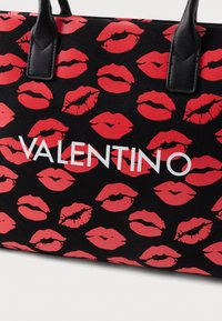 Black tote bag featuring repeated red lip prints, with white "VALENTINO" logo prominently displayed across the center; sturdy black handles.