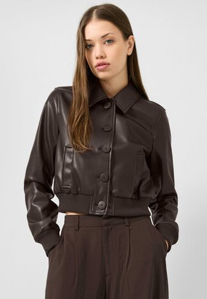 Giovane donna con lunghi capelli castani che indossa una giacca di pelle marrone scuro con bottoni e pantaloni a vita alta coordinati, con le mani nelle tasche.