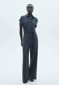 Tuta grigia scura con maniche corte, colletto a punta, bottoni frontali, vita con cintura e pantaloni a gamba larga. Tessuto di texture liscia.