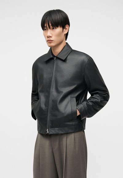 Νεαρός με ίσια μαύρα μαλλιά φοράει μαύρο δερμάτινο τζάκετ με φερμουάρ και φαρδιά καφέ παντελόνια, στέκεται με τα χέρια στις τσέπες.