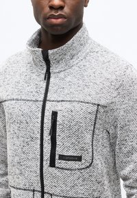 Άνδρας που φοράει γκρι υφασμάτινο μπουφάν με φερμουάρ και τσέπη στο στήθος, στέκεται μπροστά σε λευκό φόντο.