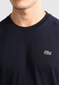 Camiseta de algodón azul marino de cuello redondo con mangas cortas y un pequeño logotipo de cocodrilo bordado en verde y blanco en el lado izquierdo del pecho.