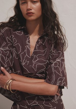 Femme aux longs cheveux foncés portant une chemise violet foncé avec des lignes abstraites blanches, les bras croisés et des bracelets dorés au poignet gauche.