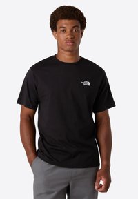 Mand med kort, krøllet hår iført en sort The North Face t-shirt og grå bukser, stående med den ene hånd i lommen mod hvid baggrund.