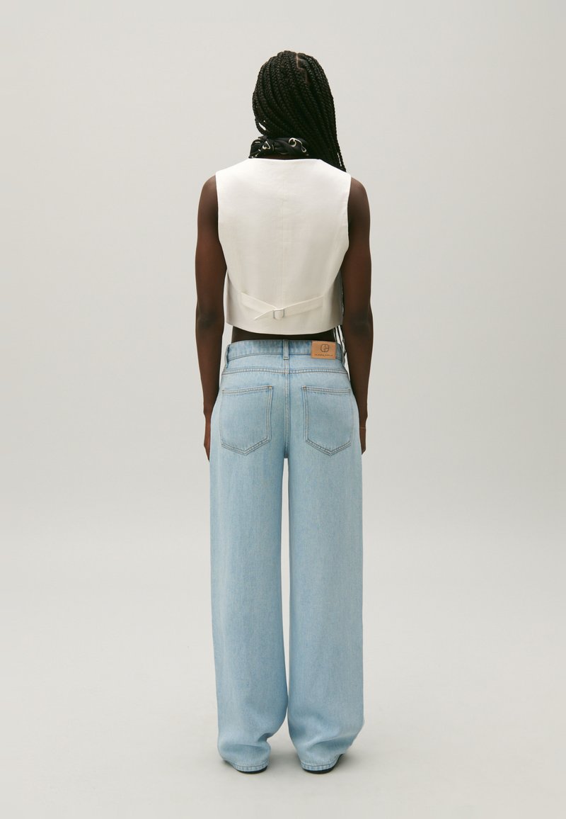 Haut blanc court sans manches avec un détail de nœud dans le dos, associé à un jean bleu clair à jambe large avec des poches arrière et une étiquette en cuir sur la taille.