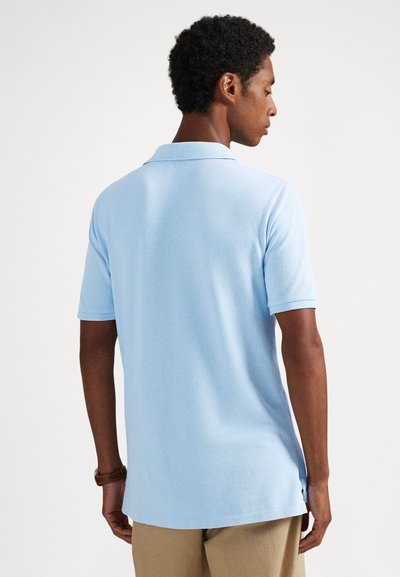 Háttal álló férfi, világoskék rövid ujjú galléros pólót és bézs nadrágot visel, egyszerű fehér háttér előtt.