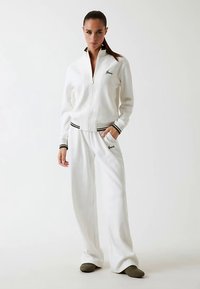 Veste blanche à fermeture éclair et pantalon assorti en tissu texturé, avec des accents rayés noirs et verts et des détails de logo brodés.