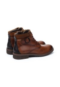 Botas de tobillo de cuero marrón con un acabado suave, tacón negro contrastante, cordones en la parte delantera y un acento decorativo de hebilla. Suela de goma plana.