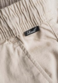 Tela beige con una cinturilla elástica fruncida, que presenta una etiqueta negra "Reell" cosida al lado. Textura suave con costuras ordenadas.
