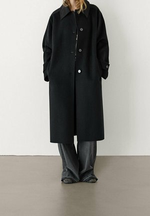 Person iført en lang sort kåbe med knapper og hænderne i lommerne, kombineret med løstsiddende mørkegrå jeans og sorte sko, stående på en ensfarvet gulv.