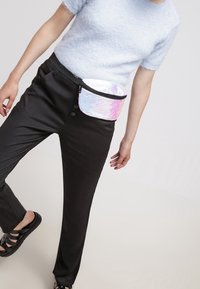 Marsupio in materiale iridescente con sfumature rosa e argento, forma rotonda, accentuato da una cerniera nera, indossato in vita sopra pantaloni neri.
