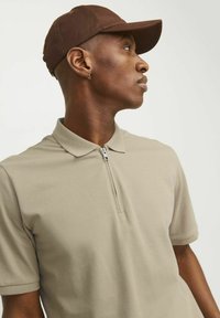 Beige polo särk, millel on tõmblukuga kaelus ja struktuurne muster, kombineerituna pruunist mütsist. Sobivus on vaba ja lõ relaxed.