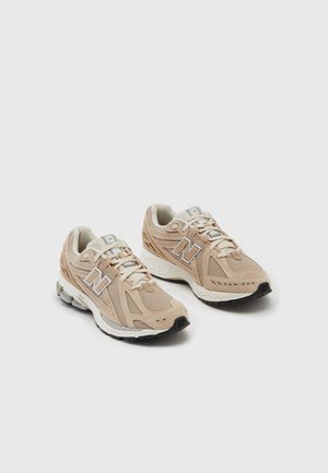 Para beżowych butów sportowych New Balance z białymi i szarymi podeszwami na jednolitym jasnym tle, ustawiona obok siebie.
