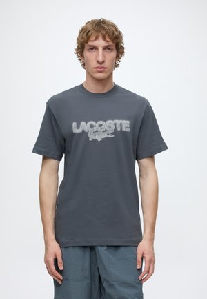 Ung mand med bølget hår iført en grå Lacoste t-shirt med hvidt logo og teksturerede blå shorts, stående foran en ensfarvet hvid baggrund.