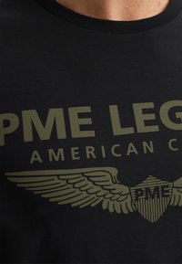 Černé tričko s velkým olivově zeleným grafickým textem "PME LEGEND AMERICAN CLASSIC" a designem křídla a štítu. Měkká bavlněná tkanina.