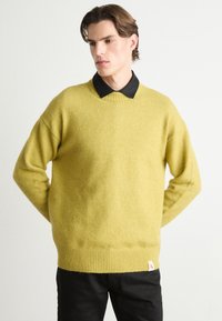 Gul ullgenser med rund hals, ribbet kant og senkede skuldre, båret over en svart skjorte med krage; har et logo-merke ved kanten.