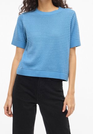 Kvinde iført en lyseblå tekstureret kortærmet sweater og sorte bukser med høj talje, stående med armene afslappet ned langs siderne.