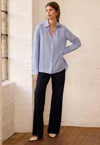 Mėlynos ir baltos dryžuotos marškiniai su krūtinės kišene, sagų užsegimu priekyje, derinami su tamsiomis kelnėmis ir smėlio spalvos batais; lygus audinys ir laisvas kirpimas.