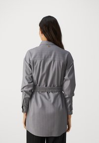 Chemise grise à manches longues avec un col, taille élastique et un motif à fines rayures, comportant un logo sur le dos.