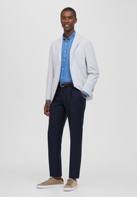 Blazer grigio chiaro, camicia blu con bottoni, pantaloni navy scuri e sneakers beige. Vestibilità slim, texture liscia, con tasche laterali e cuciture a vista.