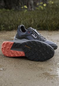 Szare buty sportowe z teksturowaną cholewką, czarną gumową podeszwą i czerwonymi akcentami. Posiadają wytrzymały bieżnik zapewniający przyczepność oraz wzmocniony pięty.