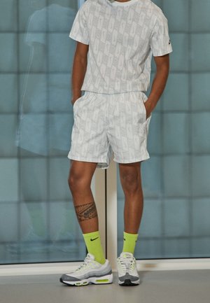 Mand iført et matchende hvidt Nike t-shirt og shorts sæt med neongule Nike sokker og hvide-grå sneakers, stående indendørs.