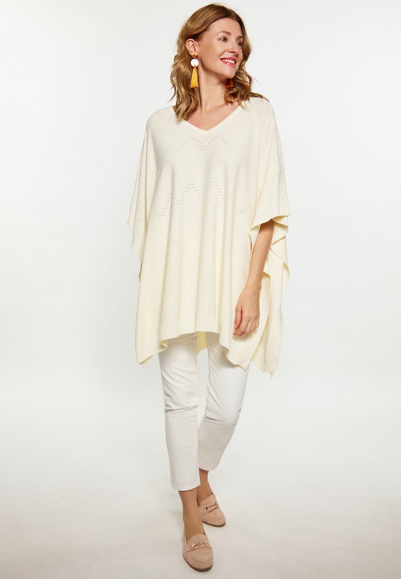 Poncho tricotat de culoare crem, cu decolteu în V, având un model de margine în zigzag. Asortat cu pantaloni albi și mocasini bej.