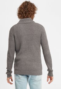 Grauer Strickpullover mit hohem Kragen, geripptem Muster, langen Ärmeln und lockerer Passform. Getragen mit hellblauen Jeans.