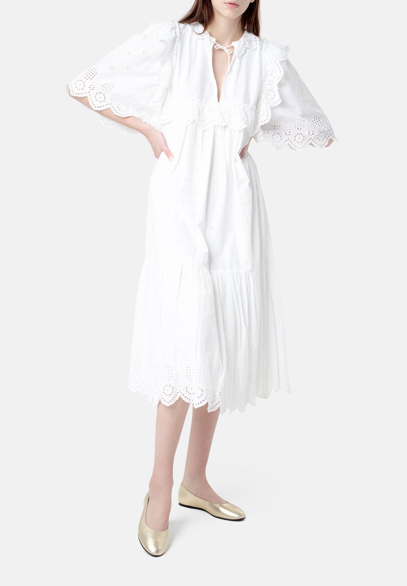 Vestido midi blanco de encaje con ojales, con un dobladillo con volantes, mangas cortas acampanadas y cierre con lazo en el cuello. Ballet flats dorados complementan el conjunto.