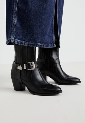 Melni ādas potītes zābaki ar smailu purngalu, kuriem ir vertikālas ribas, sudraba sprādze un grieztā papēža dizains.