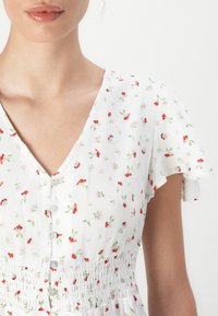 Robe blanche avec des manches courtes volantes, ornée d'un imprimé floral rouge et vert, col en V, et une taille froncée avec des détails de boutons.