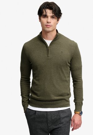 Jeune homme portant un pull zippé vert olive, un sous-vêtement blanc, un pantalon gris et une montre-bracelet, debout devant un fond uni.