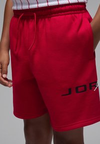 Shorts en coton rouge avec une ceinture élastique et un cordon de serrage. Présentent un logo "JORDAN" noir imprimé sur le côté gauche.