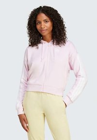 Šviesiai rožinė trumpa su kapučinu su priekine užtrauktuku; su baltais trimis dryželiais ant rankovių ir minkšta, lygi tekstūra.