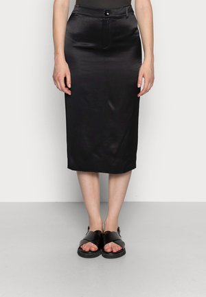 Femme portant une jupe crayon midi noire avec fermeture à boutons et à glissière, associée à des sandales noires à bout ouvert avec de larges brides croisées.