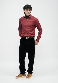 Röd långärmad skjorta, svarta manchesterbyxor, svart bälte med guldspänne, bruna sneakers. Modell poserar mot en slät vit bakgrund.