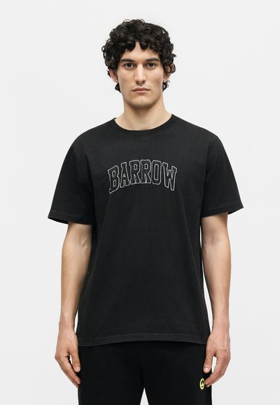 Νέος άνδρας με σγουρά μαλλιά που φοράει μαύρο T-shirt "BARROW" και μαύρο παντελόνι με μικρό κίτρινο λογότυπο με χαμογελαστό πρόσωπο.