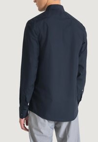 Camicia navy a maniche lunghe con colletto, polsini abbottonati, texture liscia e orlo curvo, indossata con pantaloni chiari.
