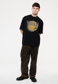 Black t-shirt with a large yellow and gray Nirvana logo. Loose fit. Paired with brown wide-leg pants and black shoes. Simple background.