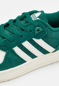 Gröna mockaskor med vita läderdetaljer, med tre vita ränder, gröna snören och en texturerad vit gummisula.