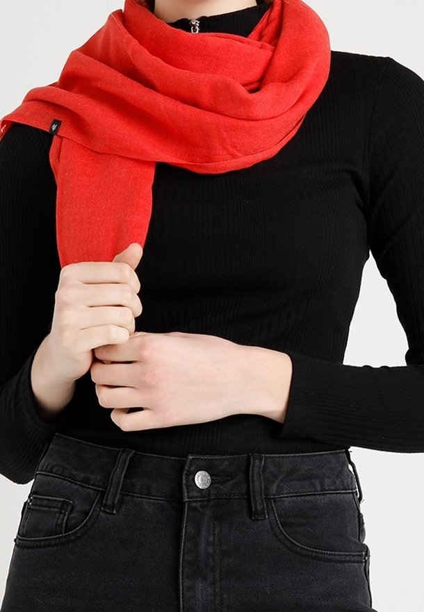 Personne portant un haut côtelé noir et un jean foncé tenant une écharpe rouge vif enroulée autour du cou et des épaules.