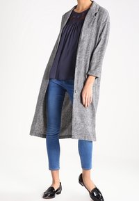 Abrigo largo de cuadros gris con un ajuste holgado, combinado con una blusa azul marino, jeans ajustados azules y mocasines negros de charol.