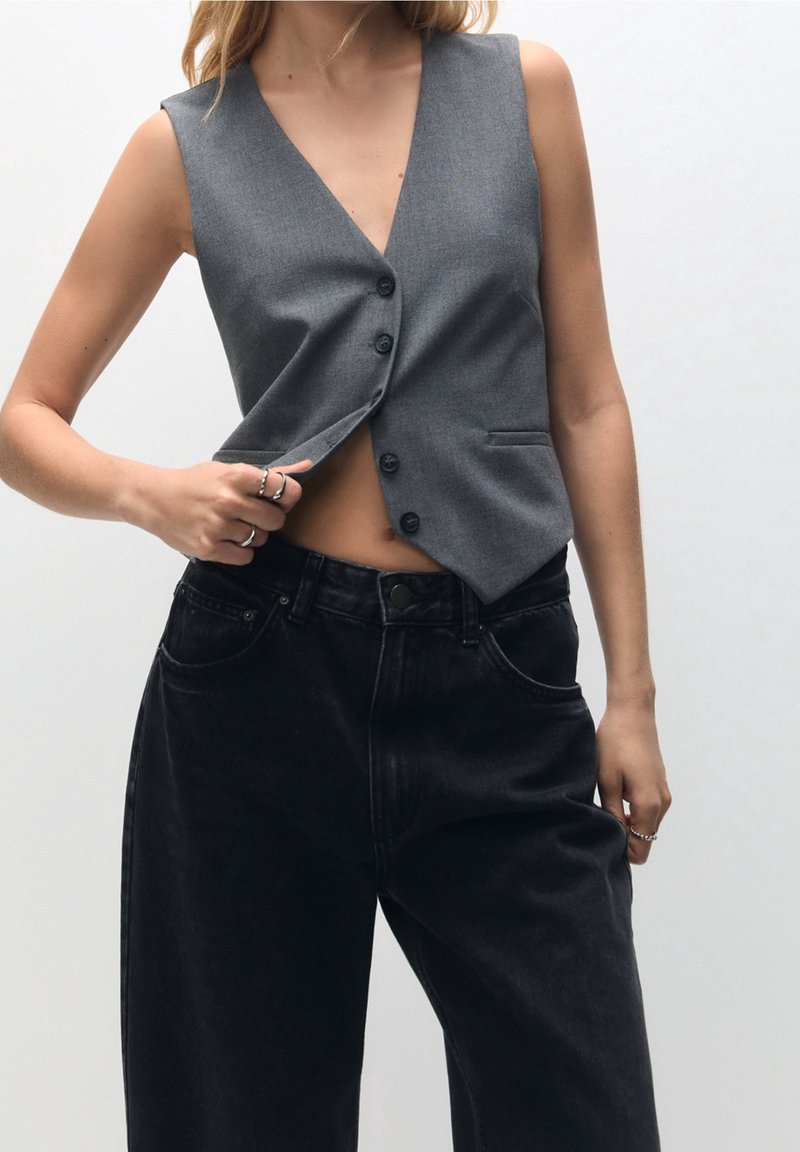 Femme portant un gilet gris sans manches boutonné et un jean noir taille haute, soulevant légèrement le gilet pour révéler la peau en dessous.